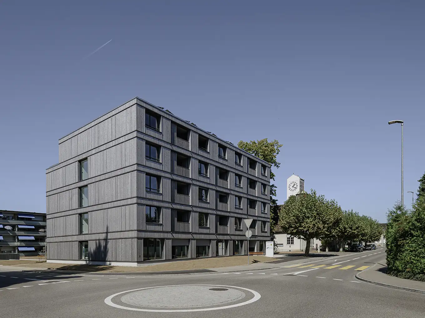 Modernes Wohngebäude mit einem Kreisverkehr in der Mitte, umgeben von Grünflächen und Parkplätzen.